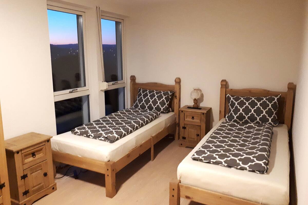 A room with two single beds, each with a wooden headboard and a patterned bedspread. There are two bedside tables with lamps, and a small wooden nightstand with drawers. The room has a window with a view of the city at dusk.