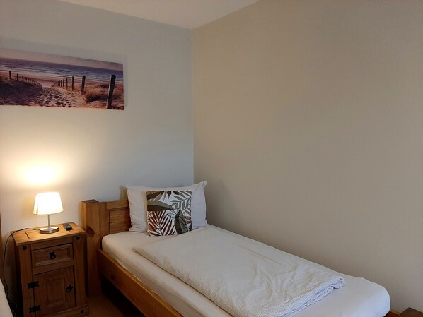 A single bed with a white mattress and a patterned pillow, a wooden nightstand with a lamp, and a wall-mounted painting of a beach scene. - Fiore Gästezimmer E11 (Esselbach)