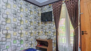 A room with a patterned wall, a bed with a colorful bedspread, a TV mounted on the wall, and a window with curtains.