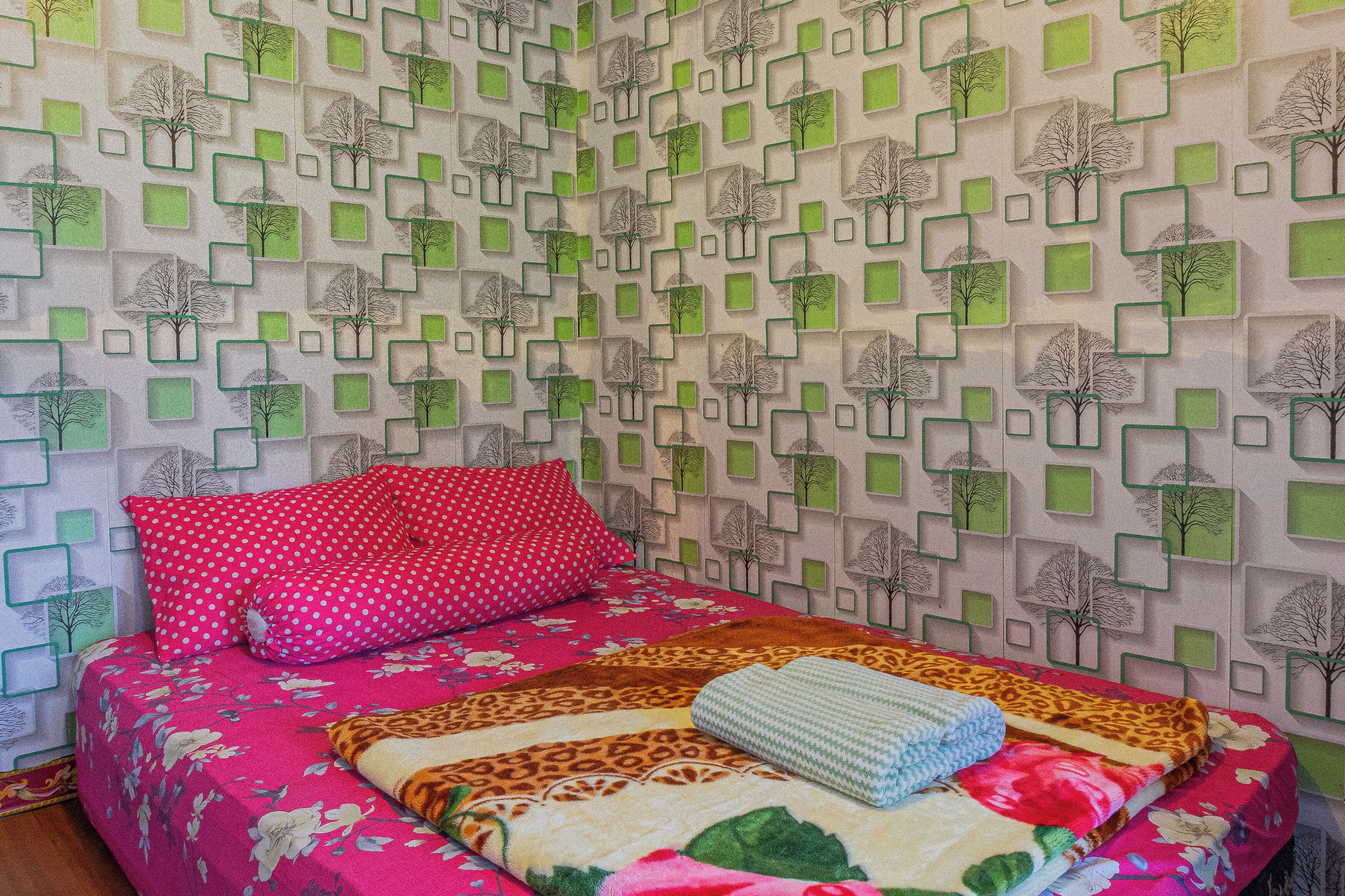 Un dormitorio con papel tapiz estampado, una cama con colcha floral y un cojín rojo con lunares.