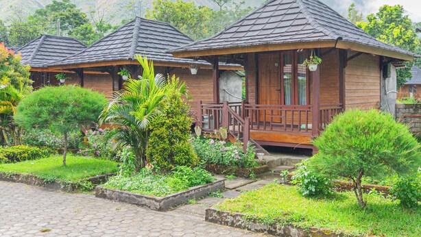 A row of wooden cabins with tiled roofs, surrounded by lush greenery and a mountainous backdrop.