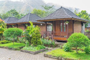 A row of wooden cabins with tiled roofs, surrounded by lush greenery and a mountainous backdrop. - Puncak Rinjani Lodge (Sembalun Lawang)