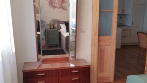 Una habitación con un aparador de madera, un otomano verde, un espejo y una cocina visible a través de una puerta abierta.