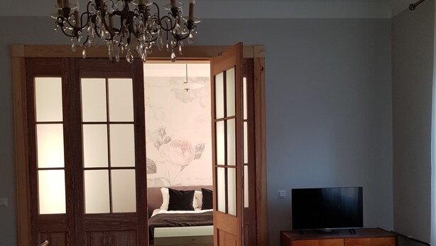 Una sala de estar con piso de madera, un sofá, un televisor sobre un mueble de madera, una araña y una puerta que da a otra habitación.