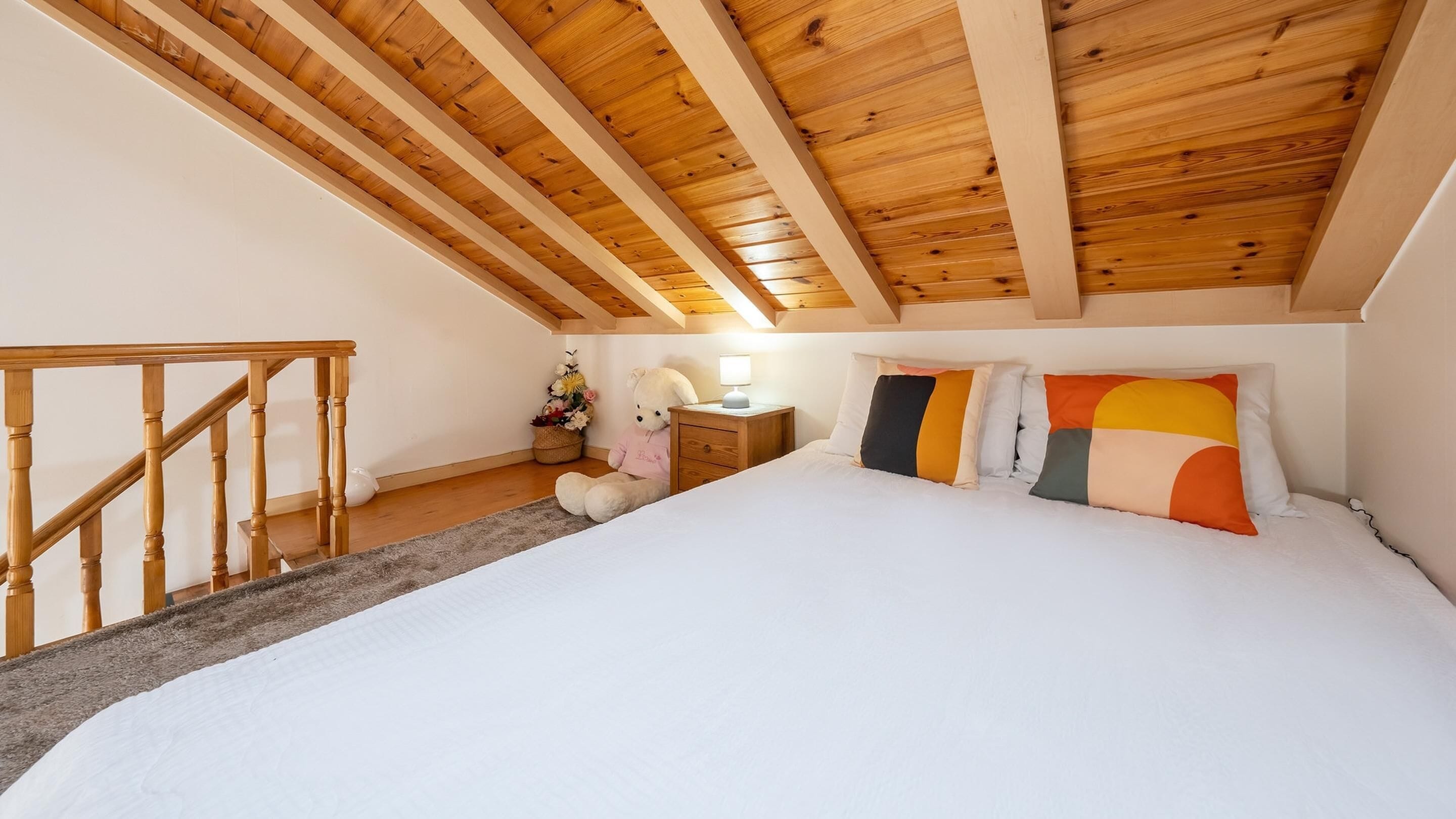 A bedroom with a wooden ceiling, a bed with colorful pillows, a nightstand, a teddy bear, and a staircase.