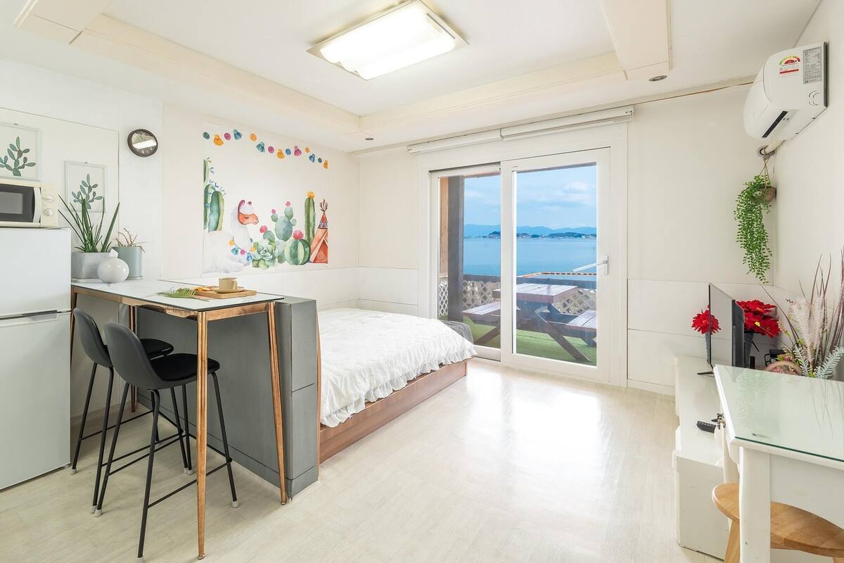 A compact bedroom with a bed, a small desk with a chair, a refrigerator, and a large window offering a view of the sea.