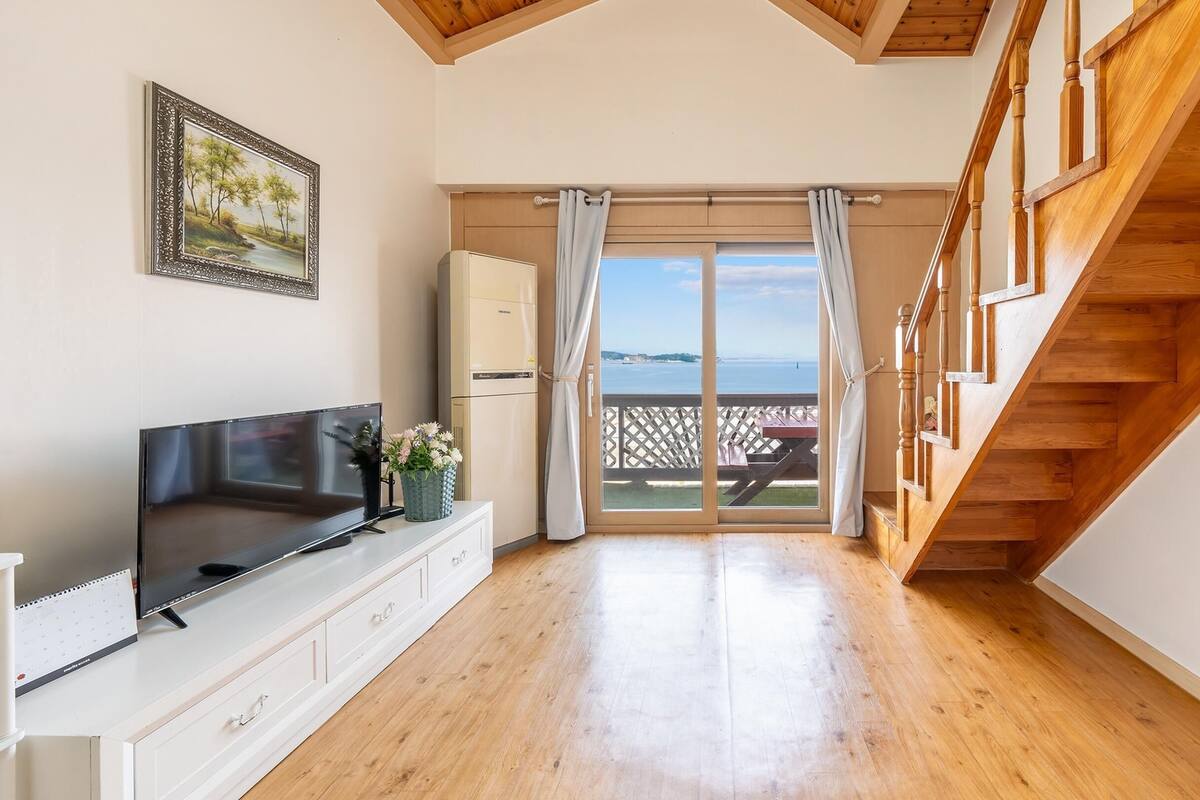 A spacious room with a wooden staircase, a large TV, and a view of the sea through a sliding glass door.