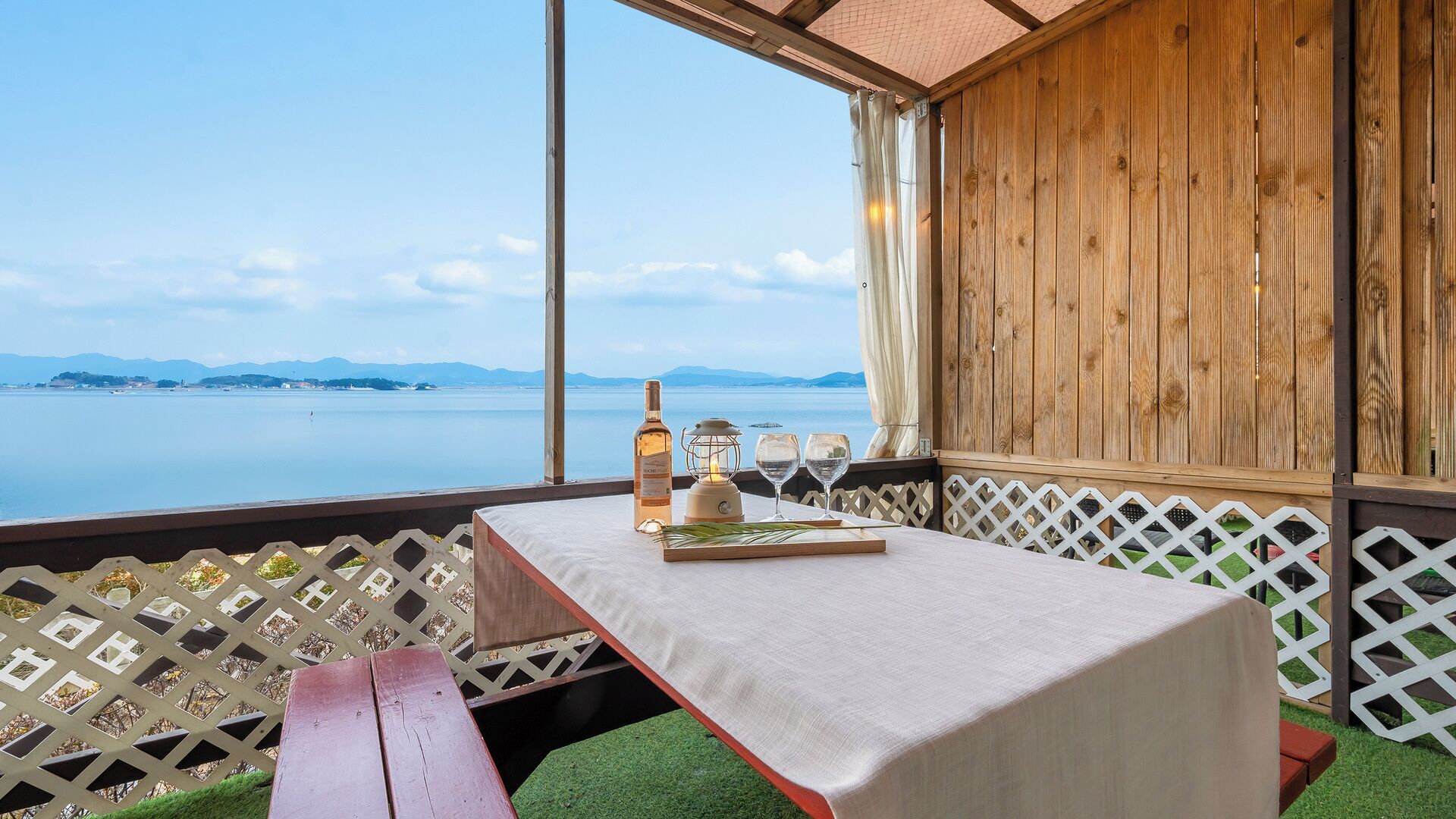 Una mesa dispuesta con una botella, vasos y un farol en un balcón con vista al mar.