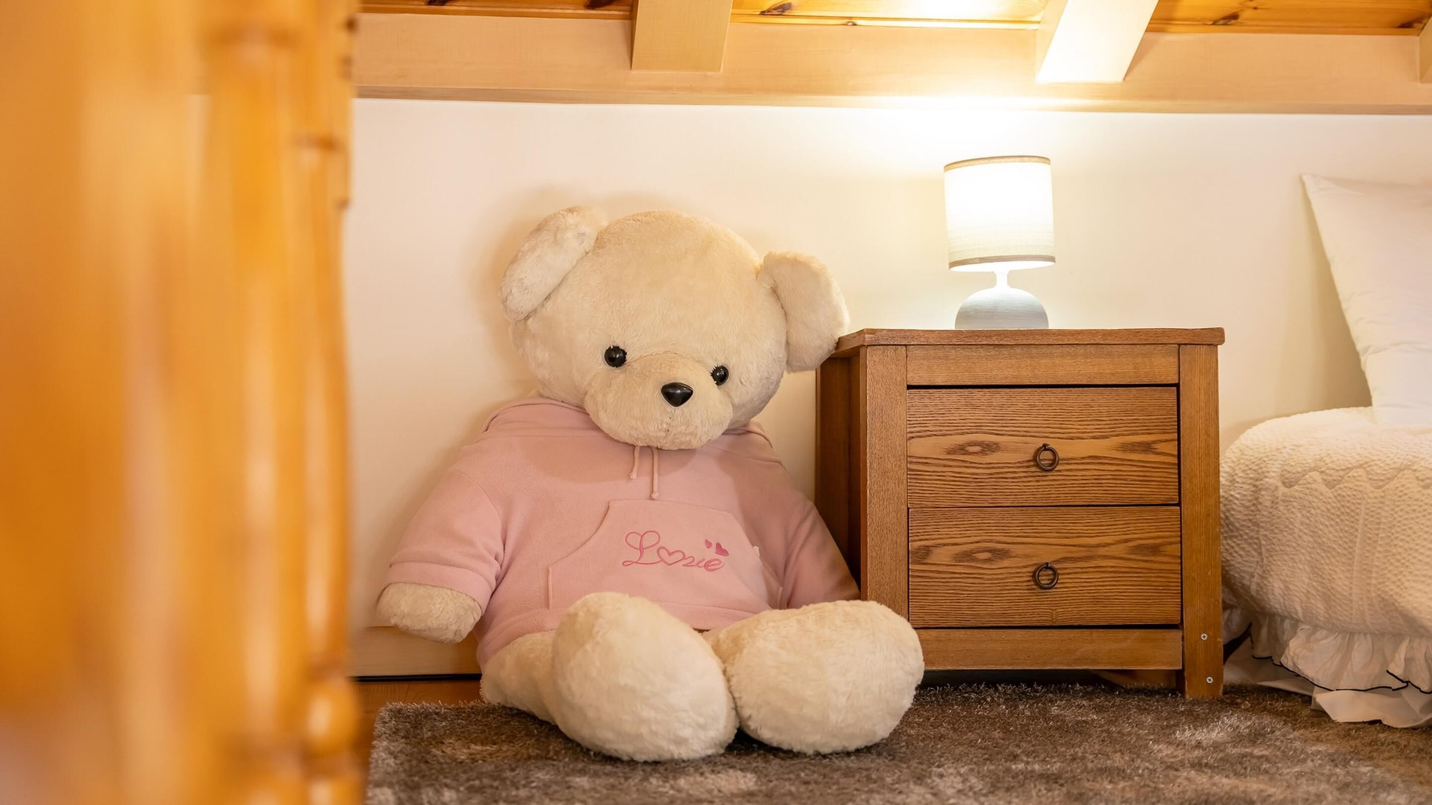 A teddy bear in a pink shirt sitting next to a wooden nightstand with a lamp.