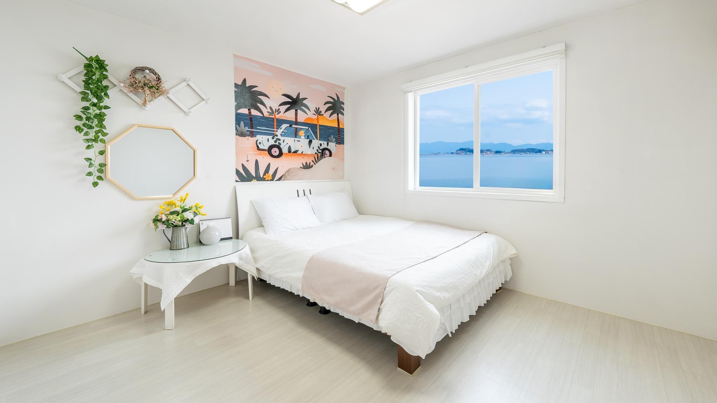 A bedroom with a bed, a bedside table with a vase of flowers, a wall-mounted mirror, and a large window with a view of the sea.