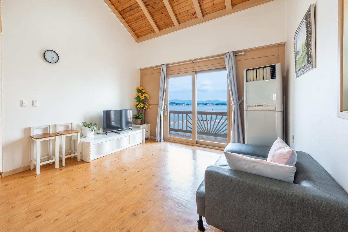 A spacious living room with a wooden floor, a grey sofa, a TV, and a balcony with a view of the sea.