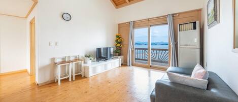 Un salón amplio con suelo de madera, un sofá gris, un televisor y un balcón con vistas al mar.