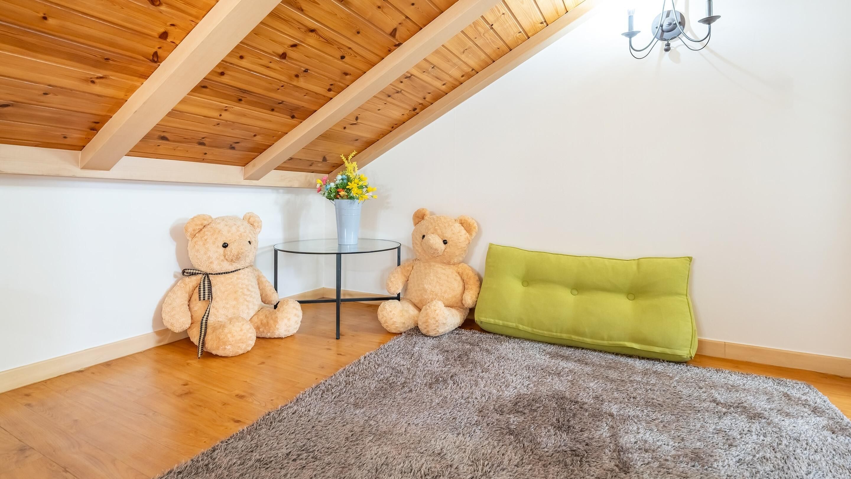 A cozy room with wooden ceiling beams, a grey shag rug, two teddy bears, a small table with a vase of flowers, and a green upholstered headboard.