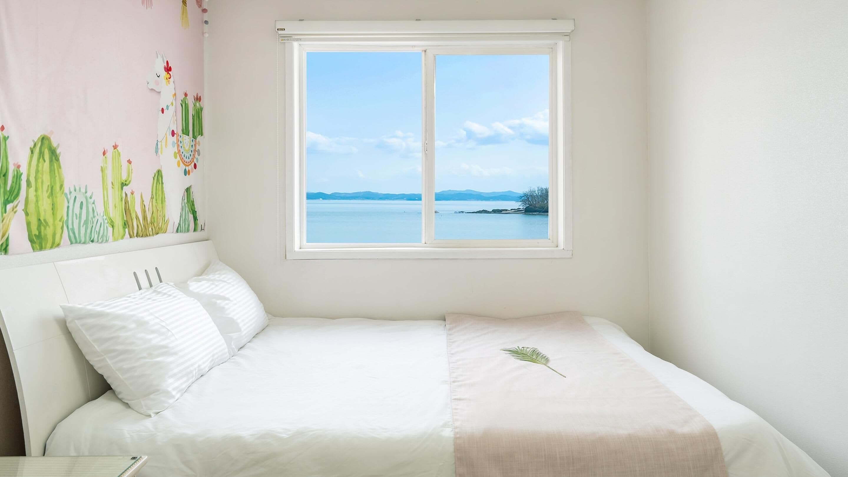 A bedroom with a bed, a window with a view of the sea, and a cactus wall decoration.