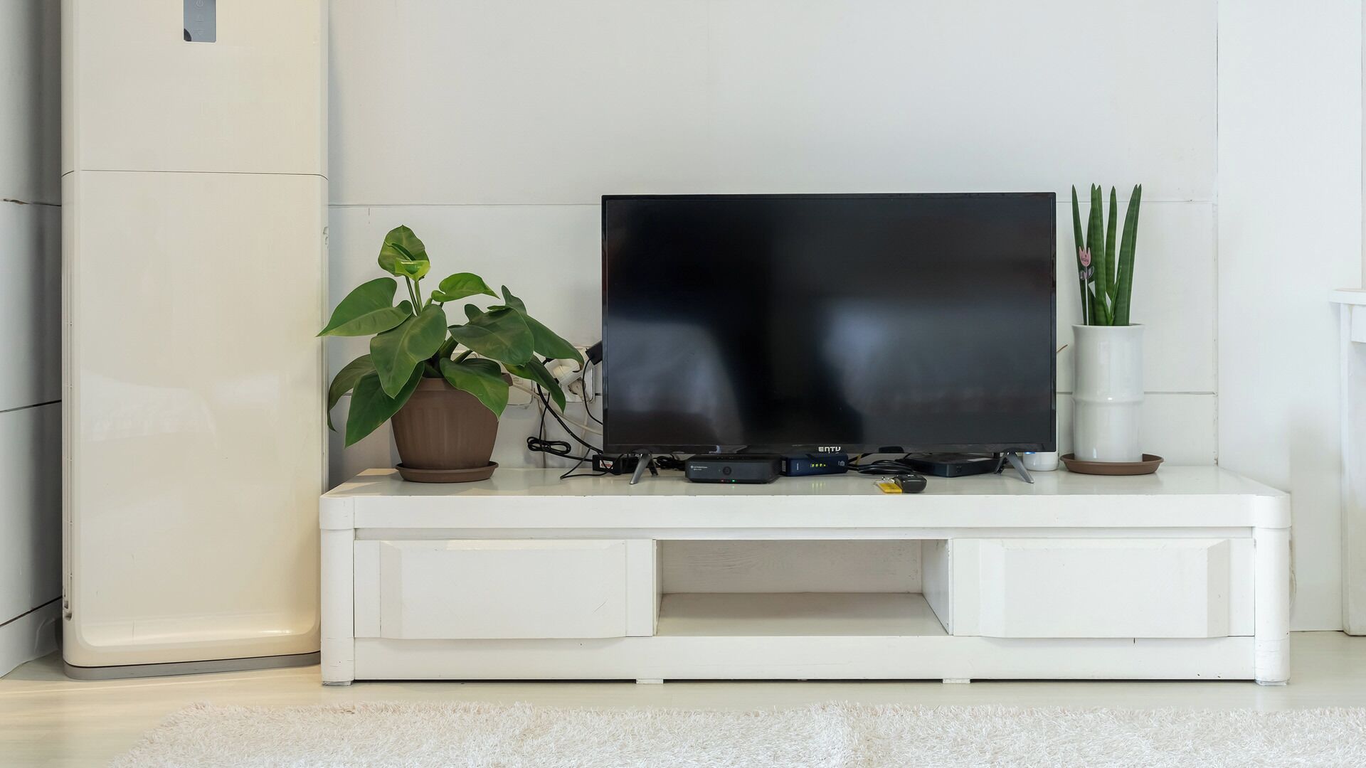 Un mueble blanco de entretenimiento con un televisor de pantalla plana, dos plantas en macetas y varios dispositivos electrónicos.