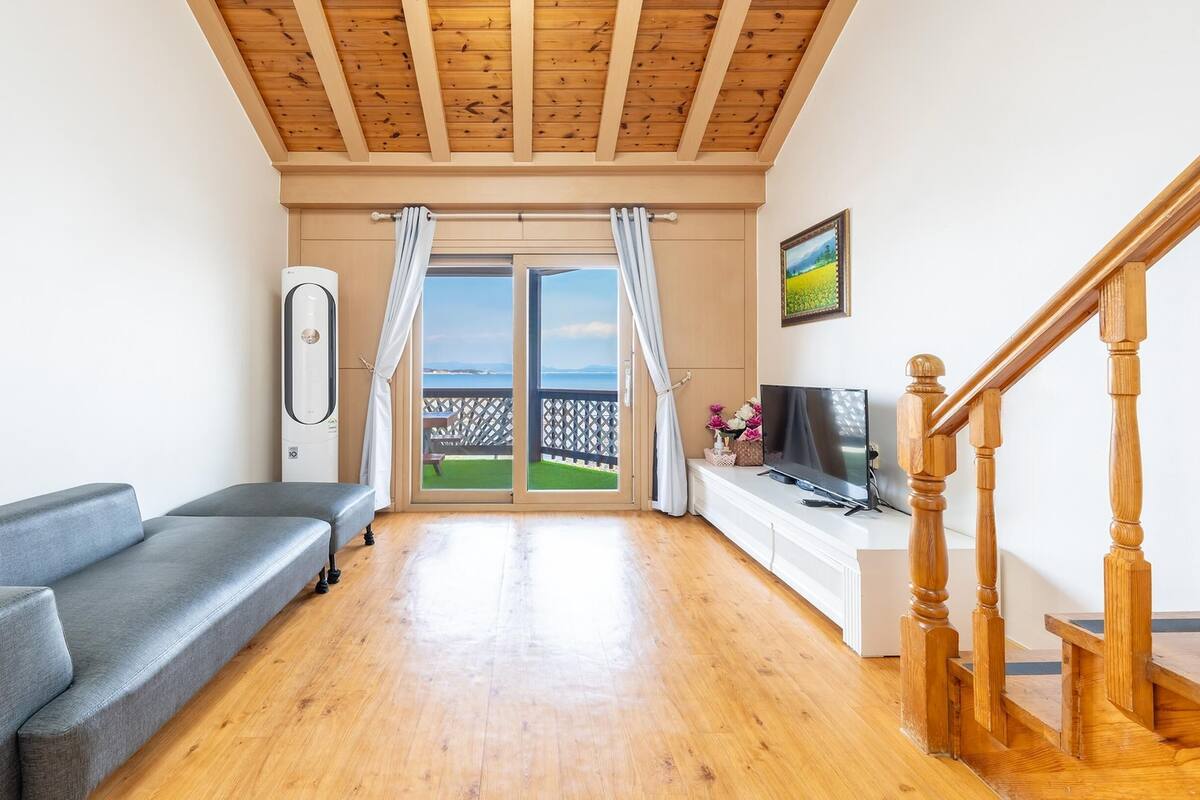 A living room with a grey sofa, wooden flooring, a TV on a white cabinet, and a wooden staircase.
