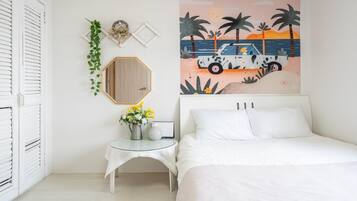 A bedroom with a bed, a small table with a vase of flowers, a wall-mounted mirror, and a beach-themed painting.