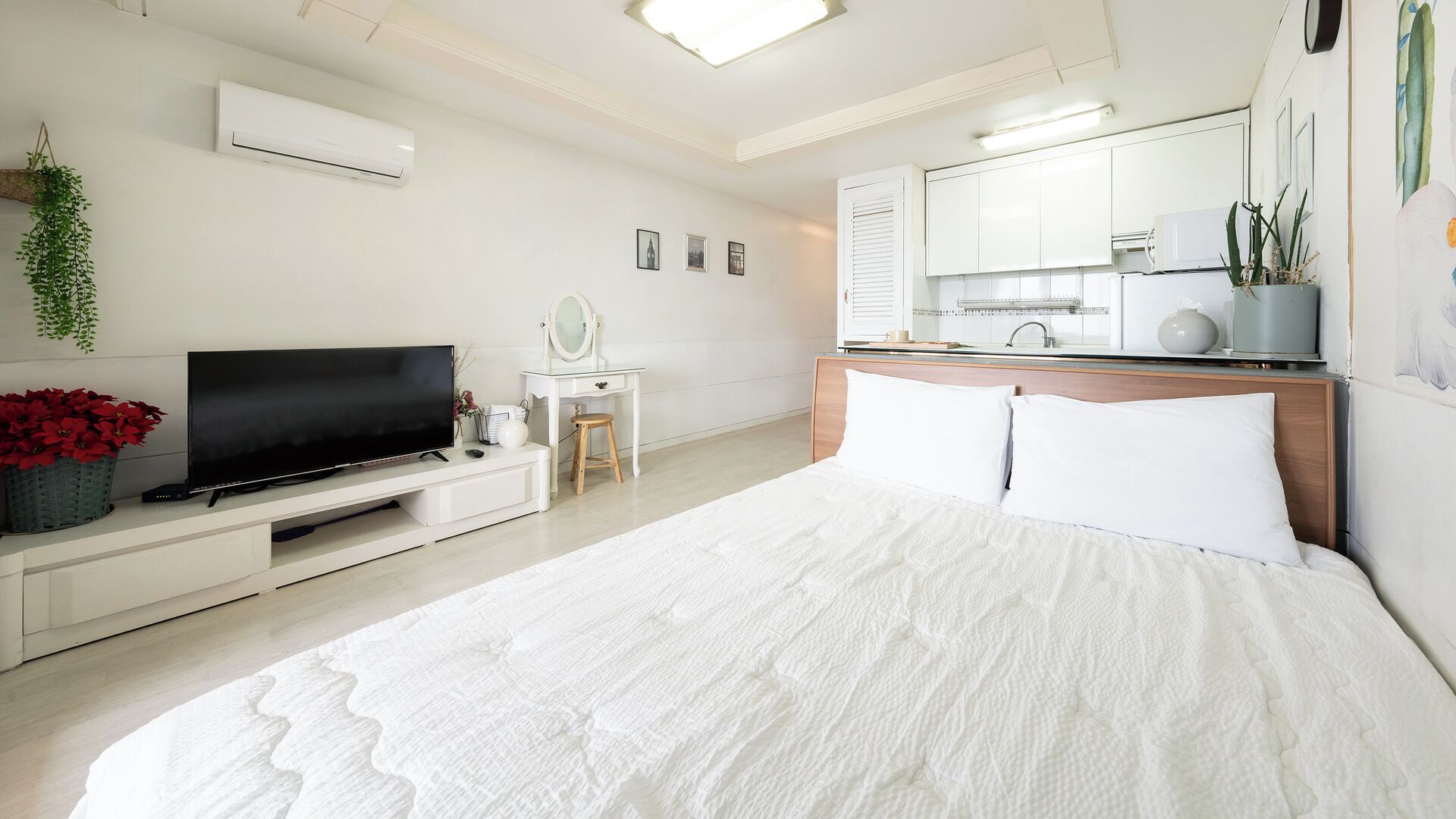 A modern bedroom with a flat-screen TV, a vanity with a mirror, a bedside table, and a bed with white bedding.