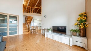 Un ampio soggiorno con pavimento in legno, una TV a schermo piatto, un tavolo da pranzo con sedie, una scala e una cucina sullo sfondo.