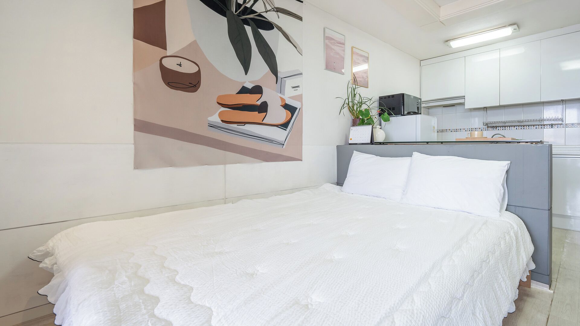 Une chambre à coucher avec un lit, une œuvre d’art accrochée au mur, une petite plante en pot et, au fond, une kitchenette.