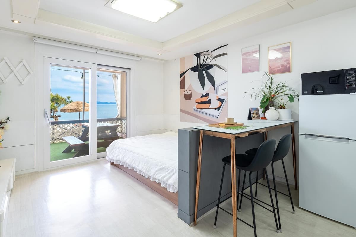 A modern, well-lit room with a bed, a small kitchenette, and a balcony with a view of the sea.