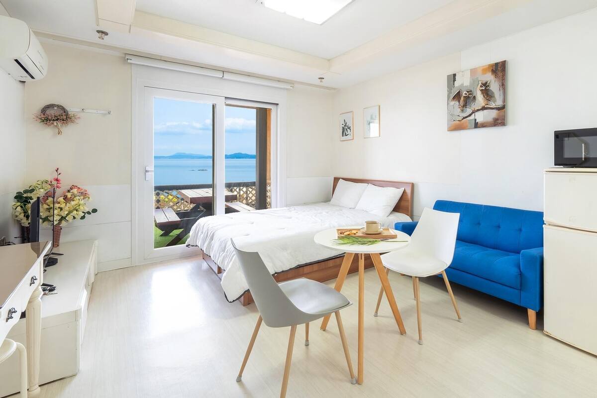 A modern bedroom with a bed, a blue sofa, a dining table with chairs, a microwave, and a view of the sea.