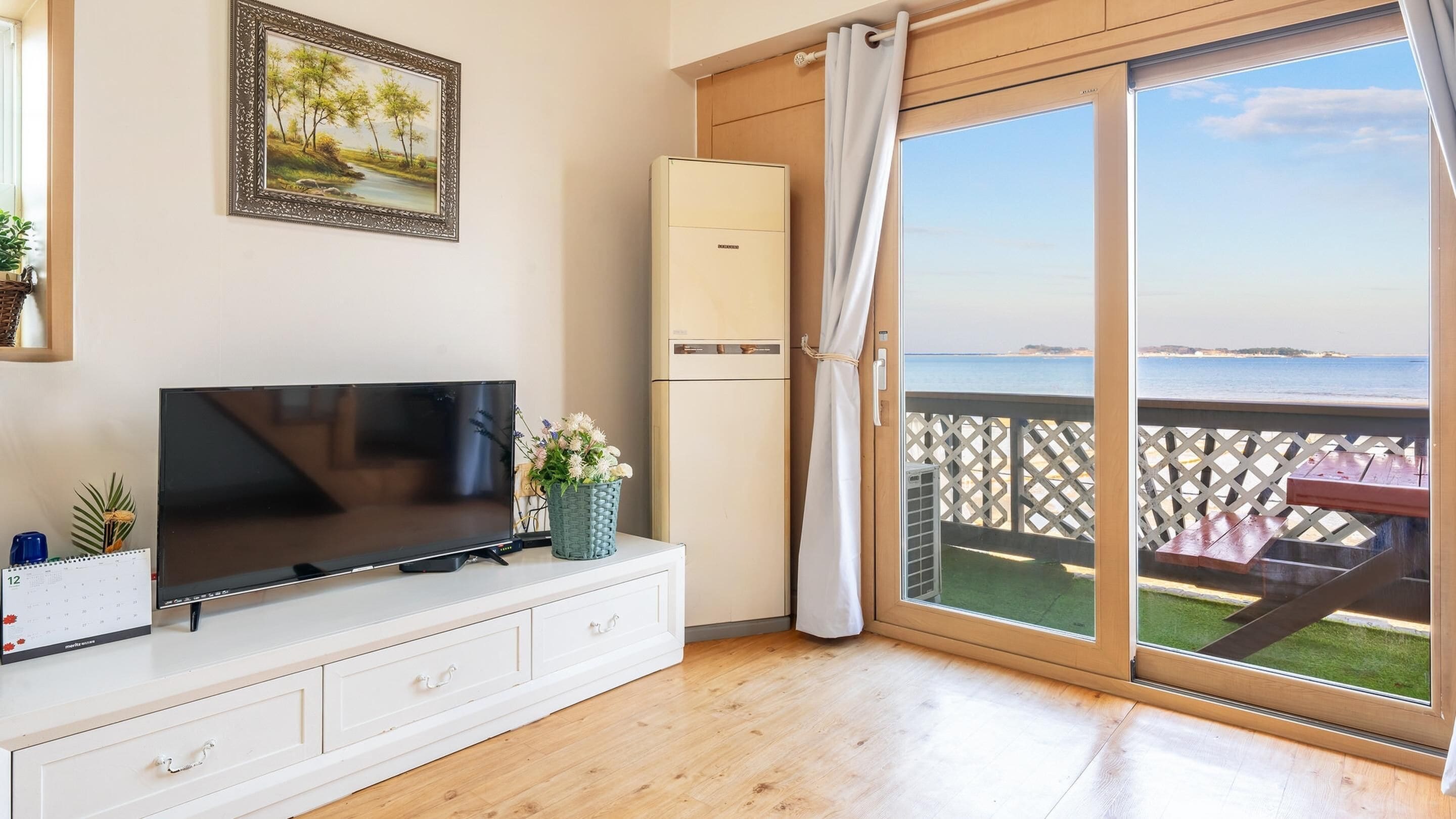 A room with a TV on a white cabinet, a calendar, a framed picture, a refrigerator, and a sliding glass door to a balcony with a view of the sea.