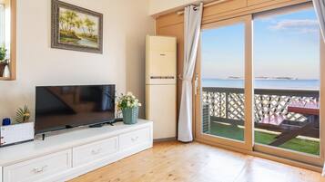 A room with a TV on a white cabinet, a calendar, a framed picture, a refrigerator, and a sliding glass door to a balcony with a view of the sea.