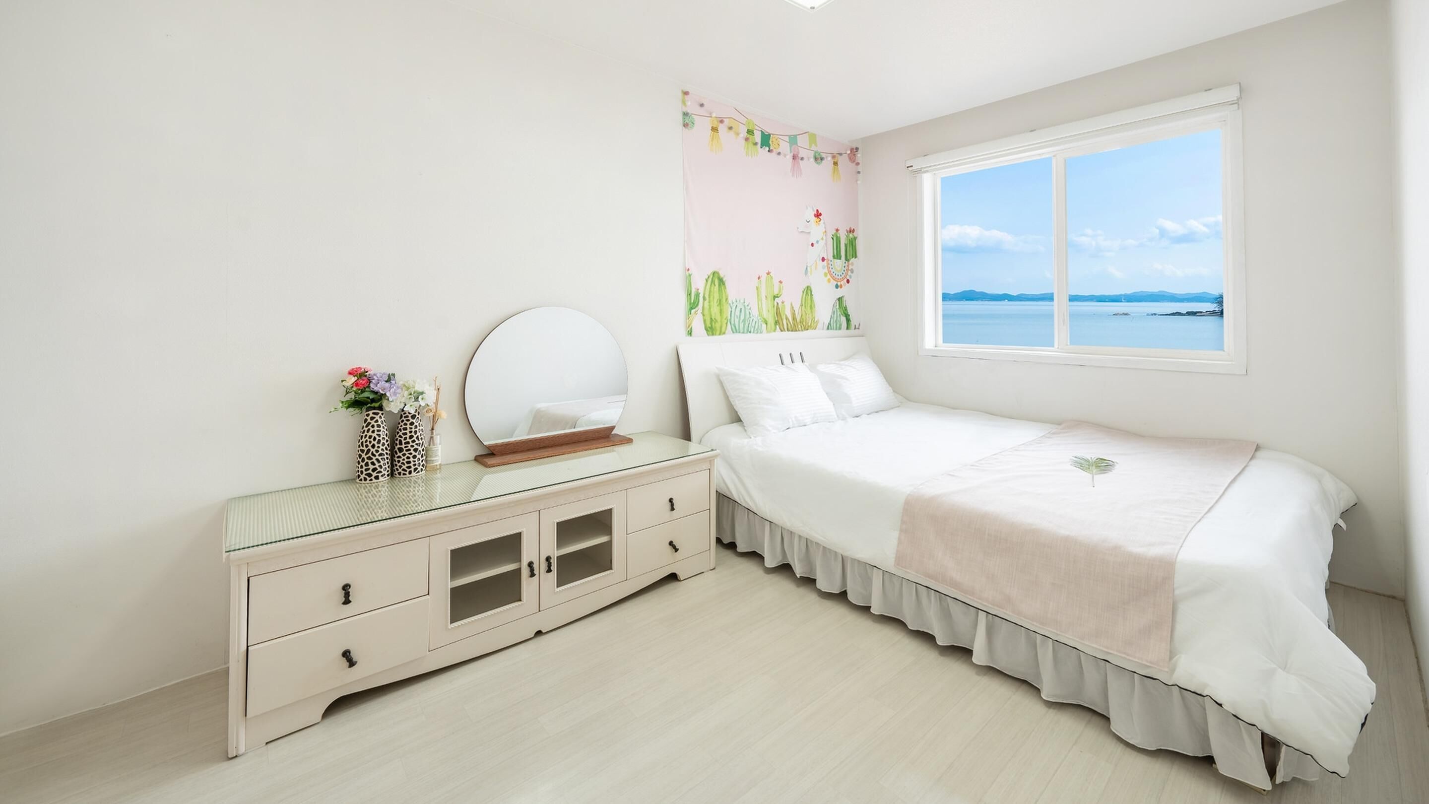 Een slaapkamer met een bed, een dressoir, een spiegel en een raam met uitzicht op zee.