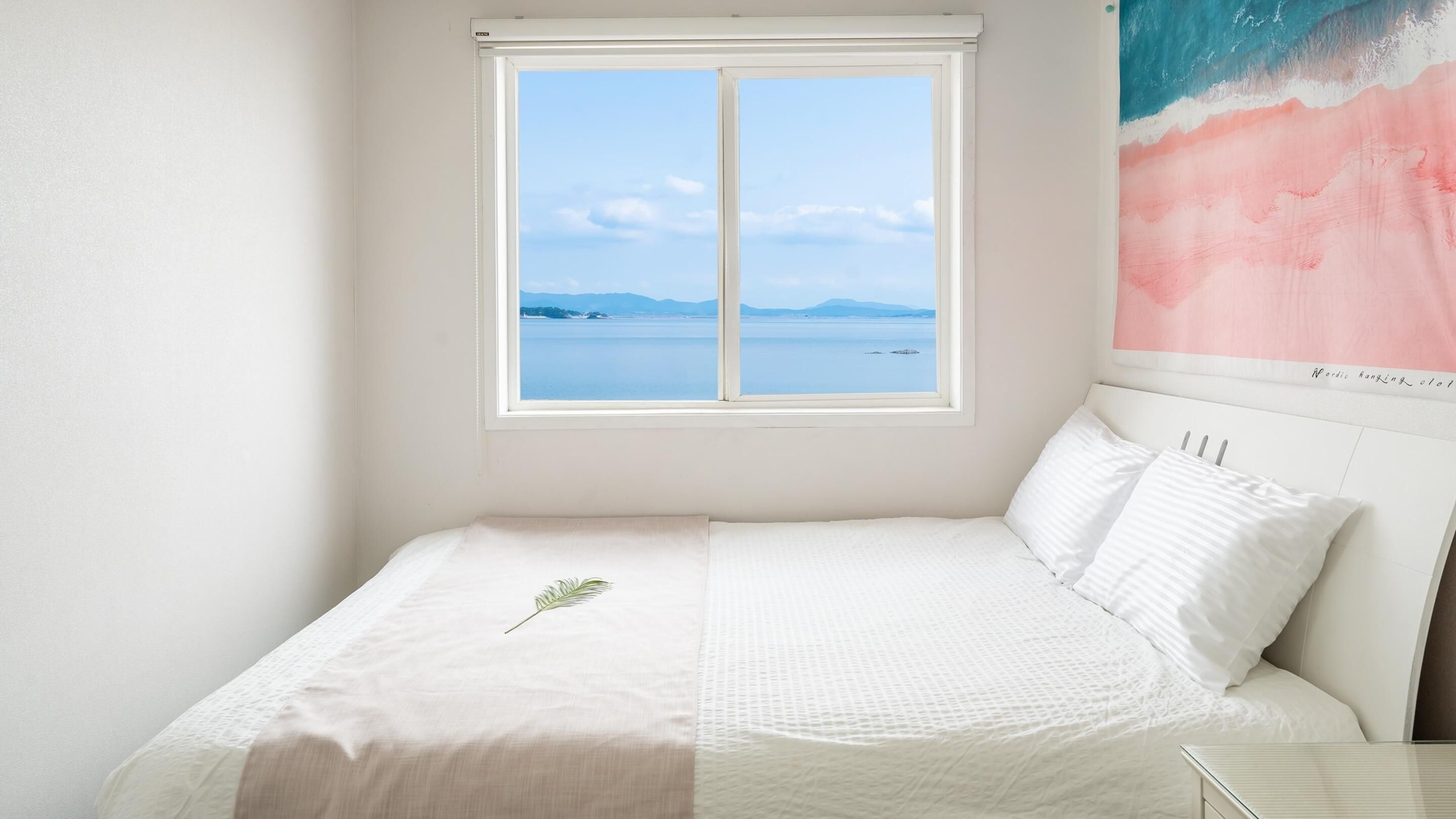 A bedroom with a bed, a window with a view of the sea, and a painting on the wall.