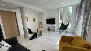 A modern living room with a sofa, a yellow armchair, a glass coffee table, a TV on a white stand, and a large window with curtains. - Bas De Villa 60m² + Jardin au Golfe de Lava (Appietto)