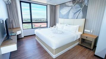 Una habitación de hotel moderna con una cama grande, un televisor sobre un mueble, una mesita de noche y un ventanal con vistas a la ciudad.