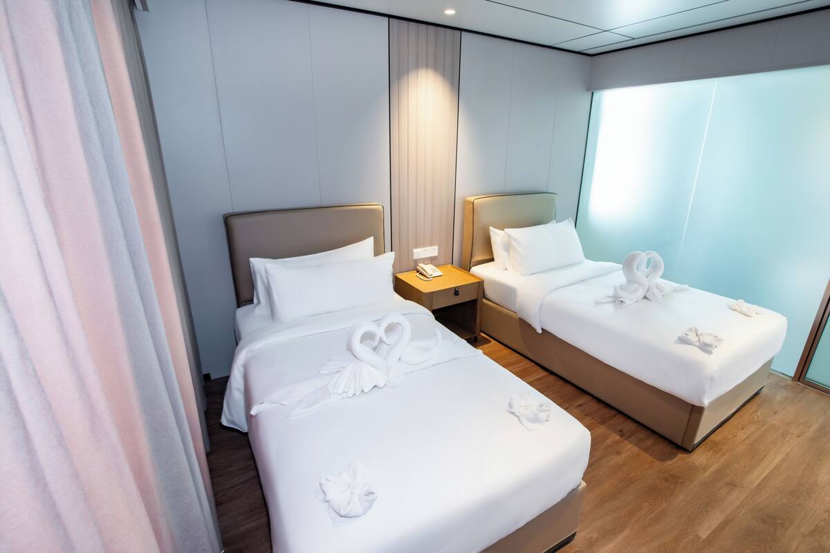 Hotelkamer met twee bedden, elk voorzien van witte lakens en decoratieve zwaan kussens. Daartussen staat een houten nachtkastje, en er is een raam met uitzicht op het exterieur.