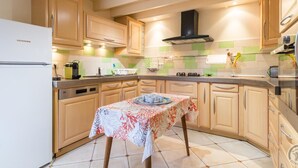 Een keuken met houten kastjes, een Liebherr koelkast, een tafel met een bloemrijk tafelkleed en een backsplash met groene en witte tegels.