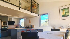 Une cuisine moderne équipée d’un four à micro-ondes, d’un four et d’une machine à café intégrés. On y trouve un coin repas avec une table et des chaises, ainsi qu’une grande fenêtre donnant sur la forêt.