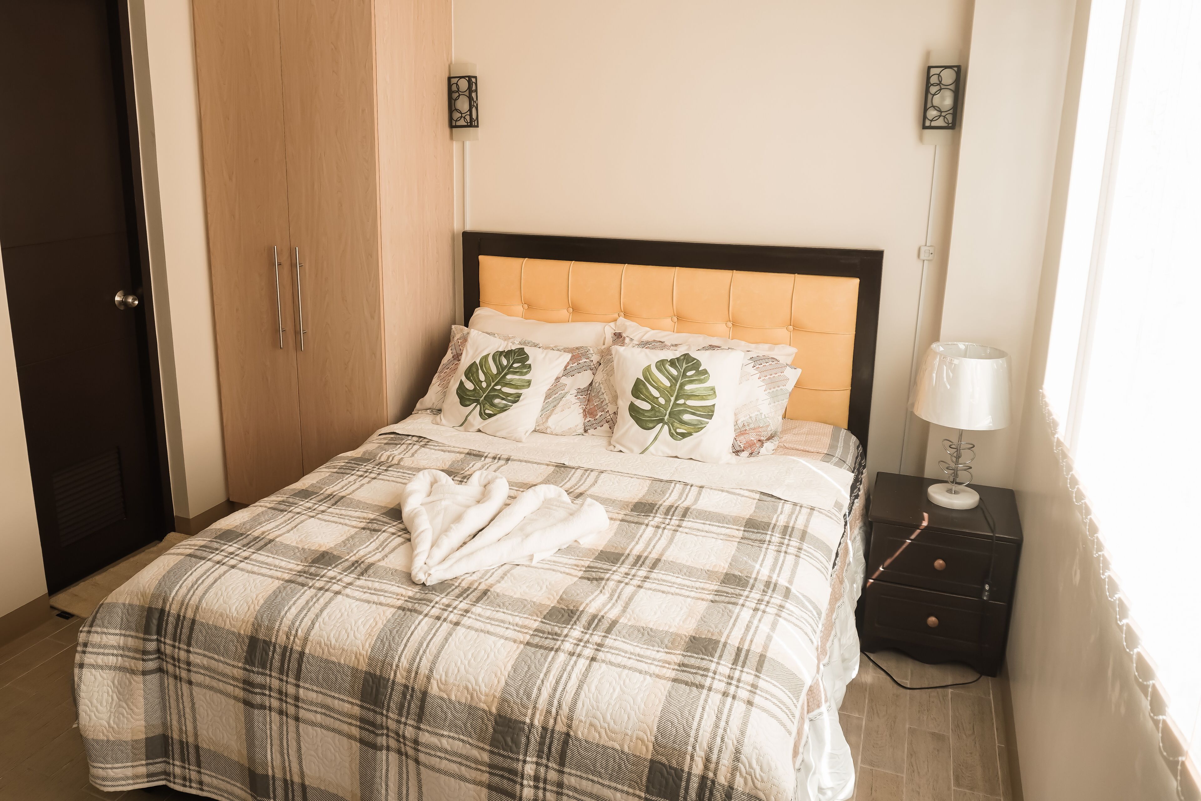 Una cama bien hecha con una colcha escocesa y dos almohadones decorativos, una mesita de noche con una lámpara y un armario con puerta.