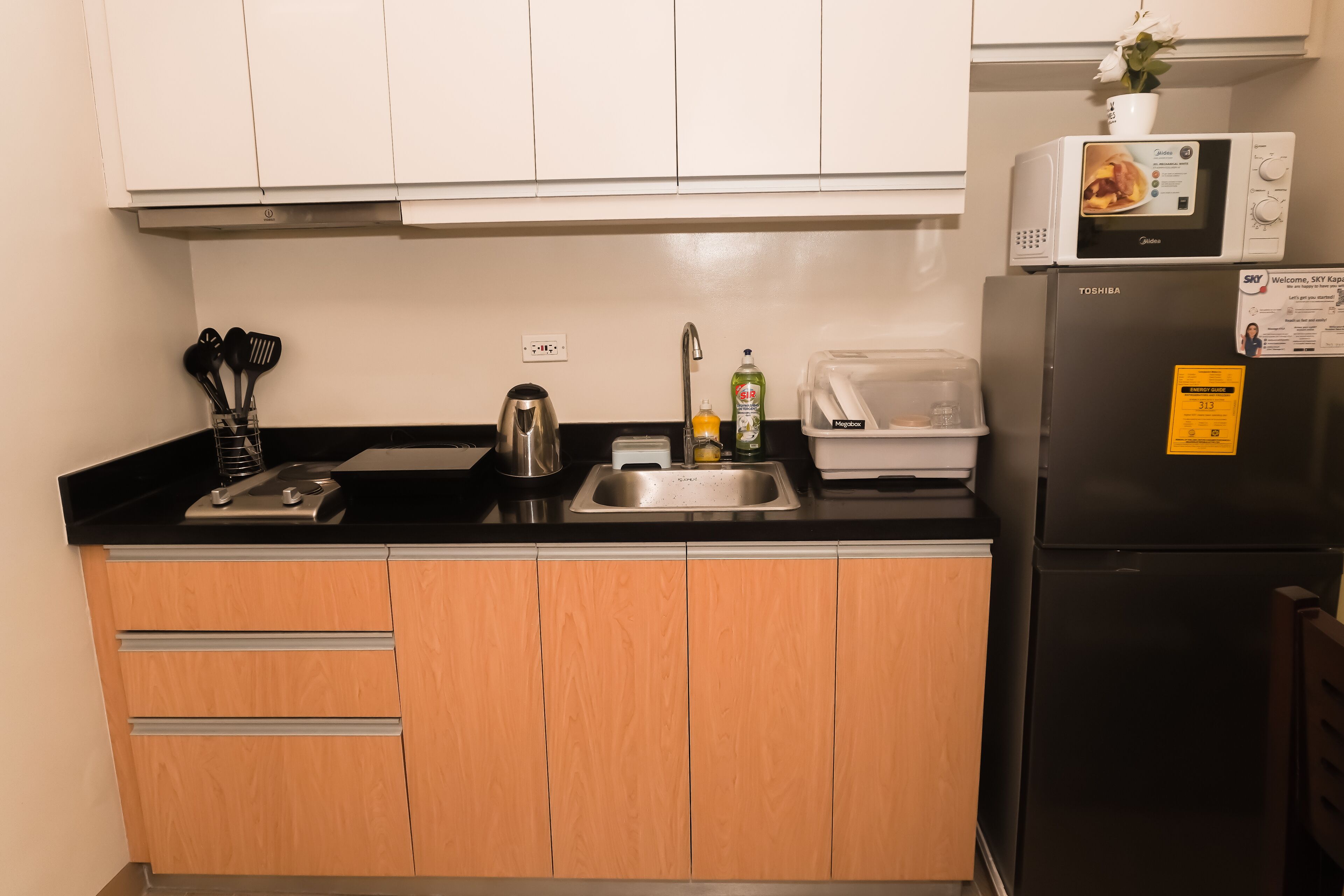 A compact kitchen with a black refrigerator, a microwave on top, a sink with a faucet, and wooden cabinets.