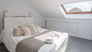 Une chambre avec un plafond en pente, un puits de lumière, un lit avec plusieurs oreillers et une couverture, et une tête de lit blanche.