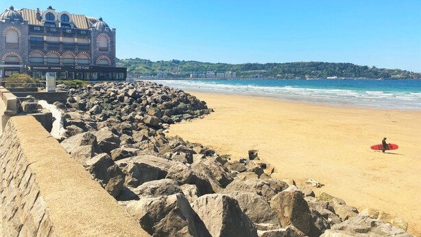 A beach with a rocky breakwater, a building with multiple floors and arched windows, and a person with a red surfboard.