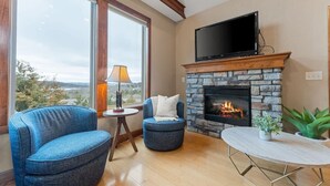 A living room with a stone fireplace, a television mounted above it, and a large window offering a view of the outdoors.