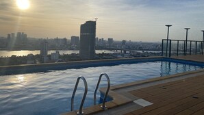 Een zwembad met metalen reling, een houten terras en uitzicht op een stadsbeeld met gebouwen en een rivier in de zonsondergang.