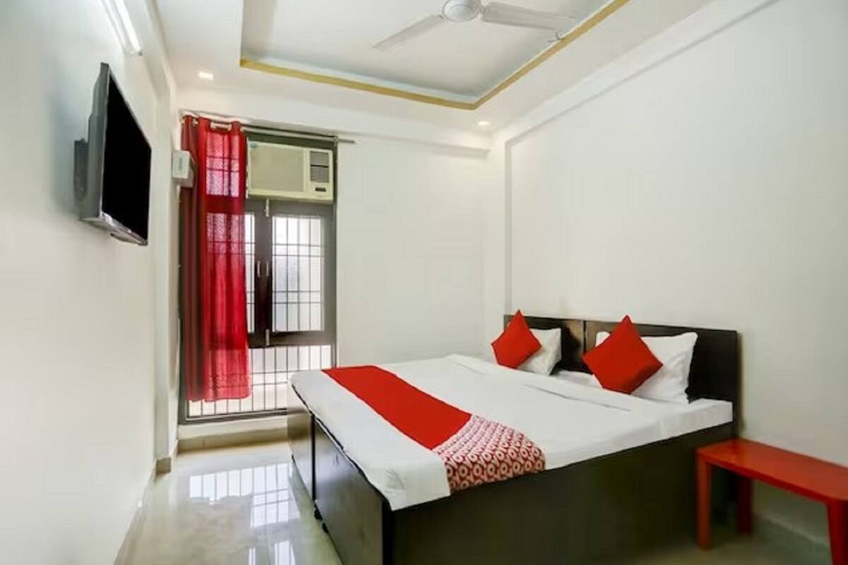 A bedroom with a bed, a TV mounted on the wall, a window with a red curtain, and a ceiling fan.