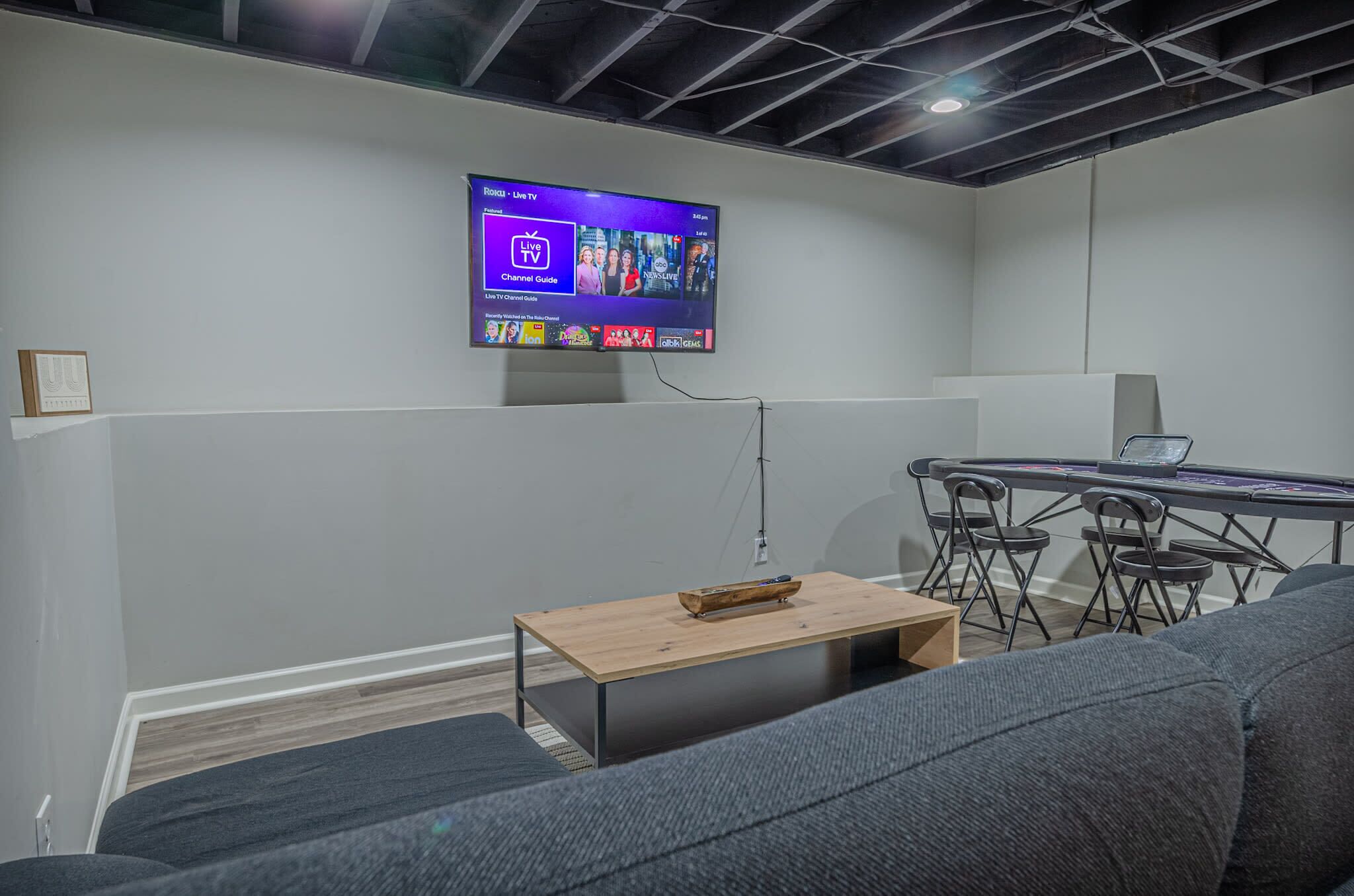 A modern office space with a mounted TV displaying a live TV channel guide, a wooden coffee table, a grey sofa, and a foldable table with chairs.