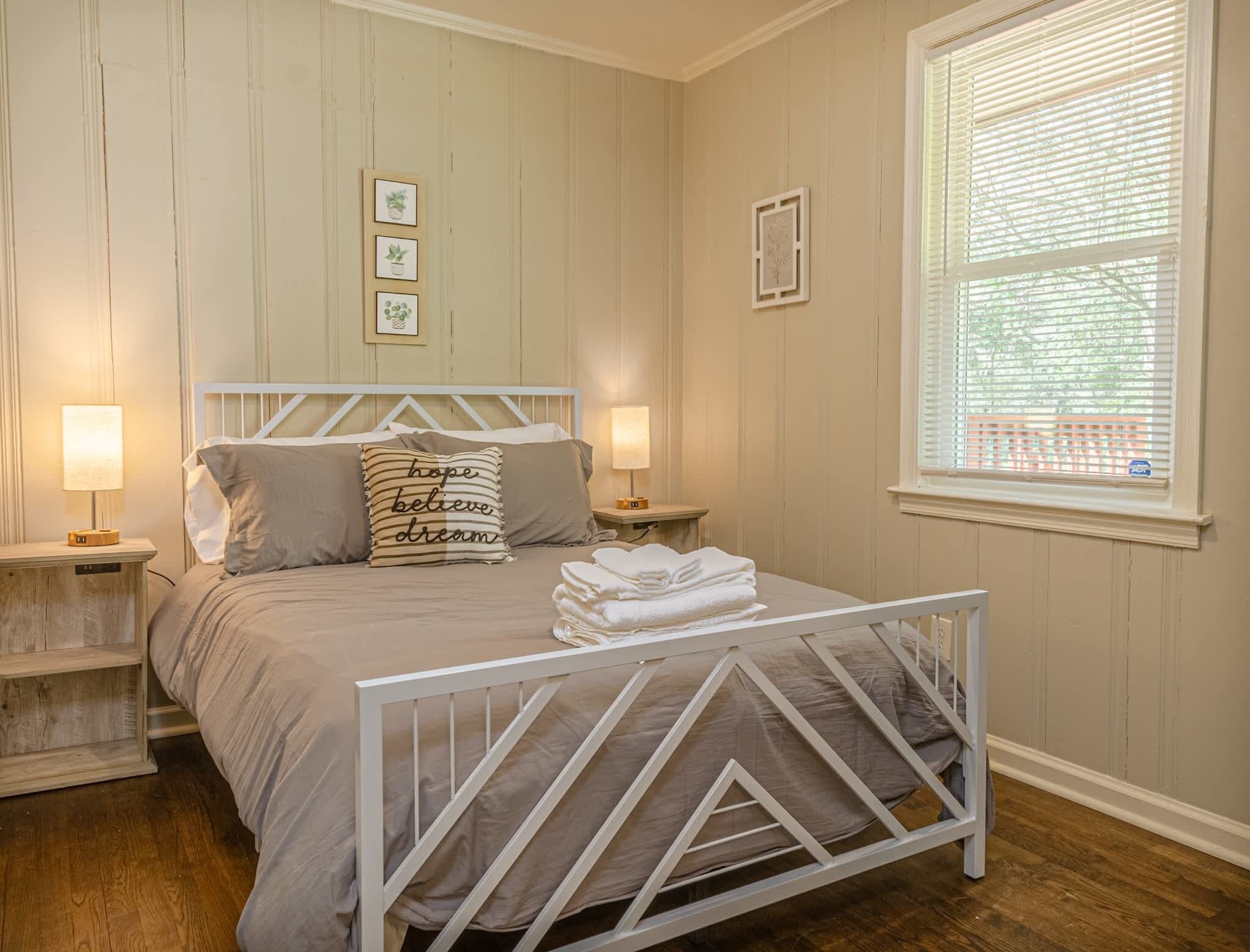 A bedroom with a bed, two bedside tables, a window with blinds, and wall-mounted artwork.