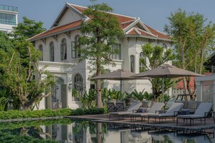 A large white building with a red-tiled roof, surrounded by lush greenery and a swimming pool with lounge chairs.