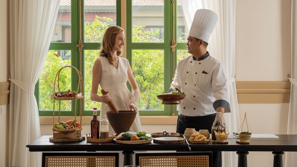 Um chef e uma mulher numa cozinha com uma janela, uma mesa com alimentos e uma tigela.