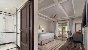 Quarto de hotel com chuveiro em gabinete de vidro, cama com tapete bege, escrivaninha com cadeira e televisão na parede.