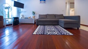 Un salon avec un canapé d’angle gris, un tapis à motifs, un ventilateur sur pied, un téléviseur fixé au mur et une fenêtre avec des rideaux.