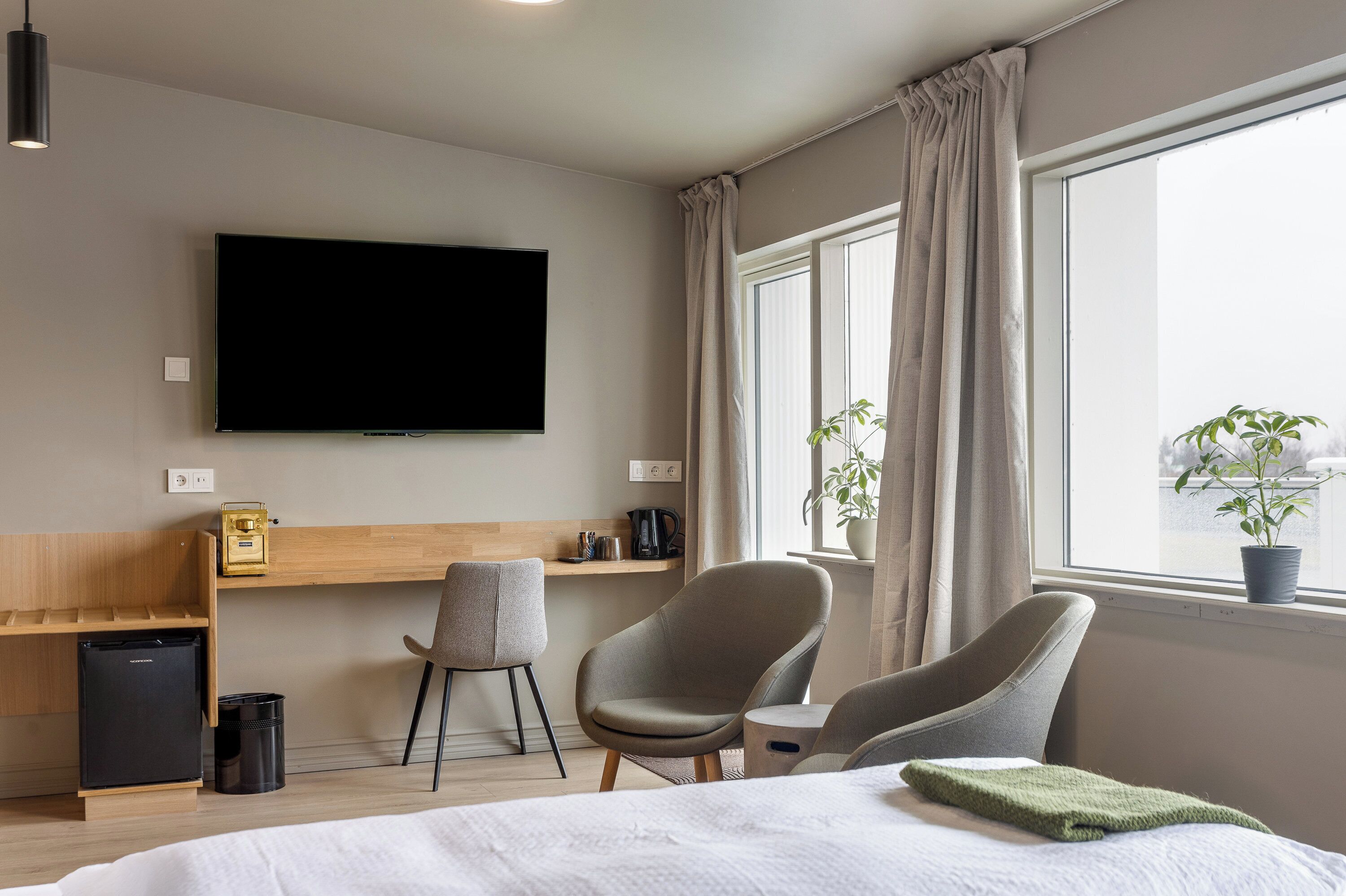 A modern hotel room with a bed, two armchairs, a desk with a TV, a small fridge, and a window with curtains.