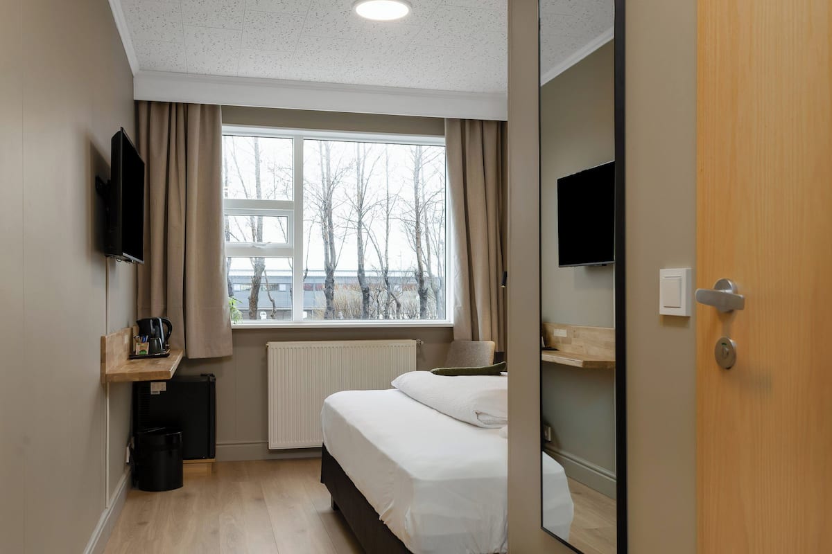 Habitación de hotel con cama, televisor montado en la pared, ventana con cortinas, radiador y un estante pequeño con cafetera.